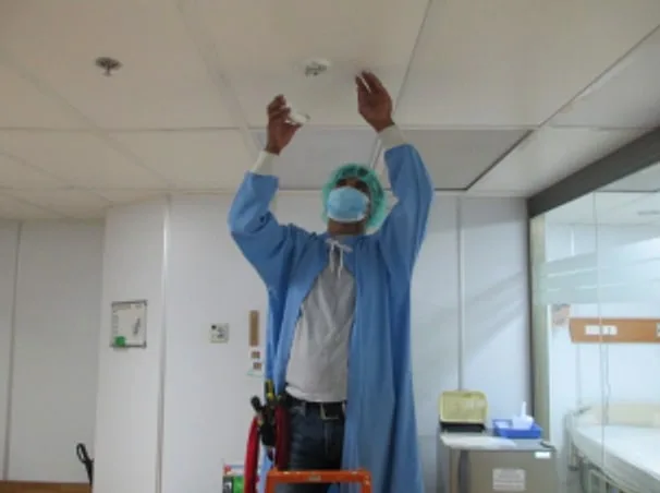 Worker in protective gown and mask reaching up to a ceiling-mounted smoke detector